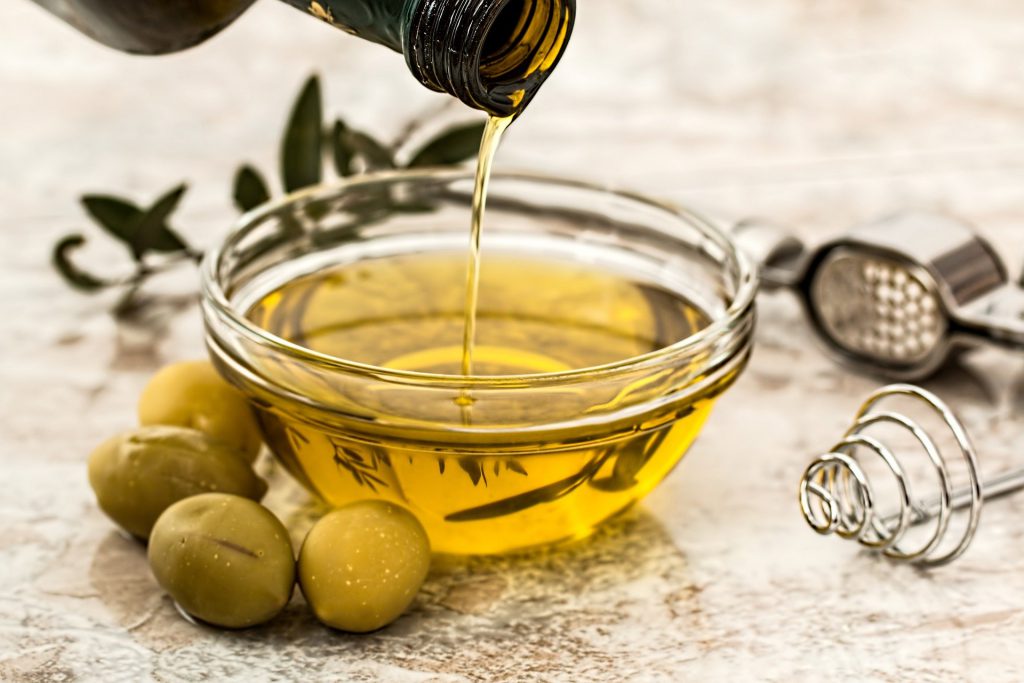 Huile d'olive versée dans un bol transparent posé à côté d'olives vertes et d'ustensiles de cuisine