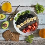 Assiette remplie de fruits posée sur table entourée par un avocat, des tranches de pain, un verre de jus d'orange et des fruits entiers